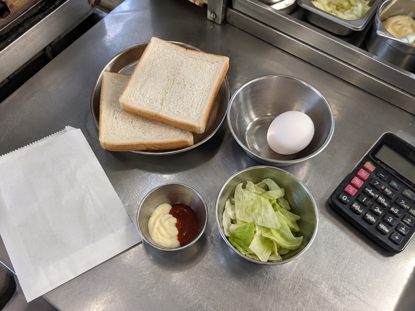 蛋吐司的麵包、蛋、蔬菜、醬料與包材分開擺放，旁邊有計算機