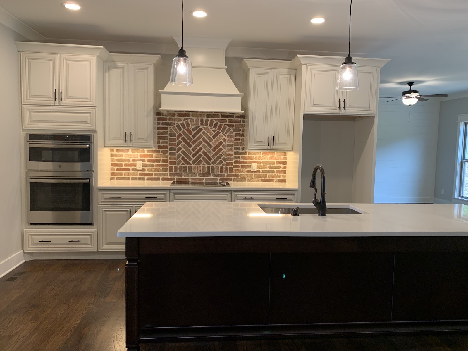Kitchen interior showing white cabinets, a double wall oven, and a brick backsplash with a herringbone pattern. A dark wood island with a white countertop sits on dark wood floors under pendant lighting.