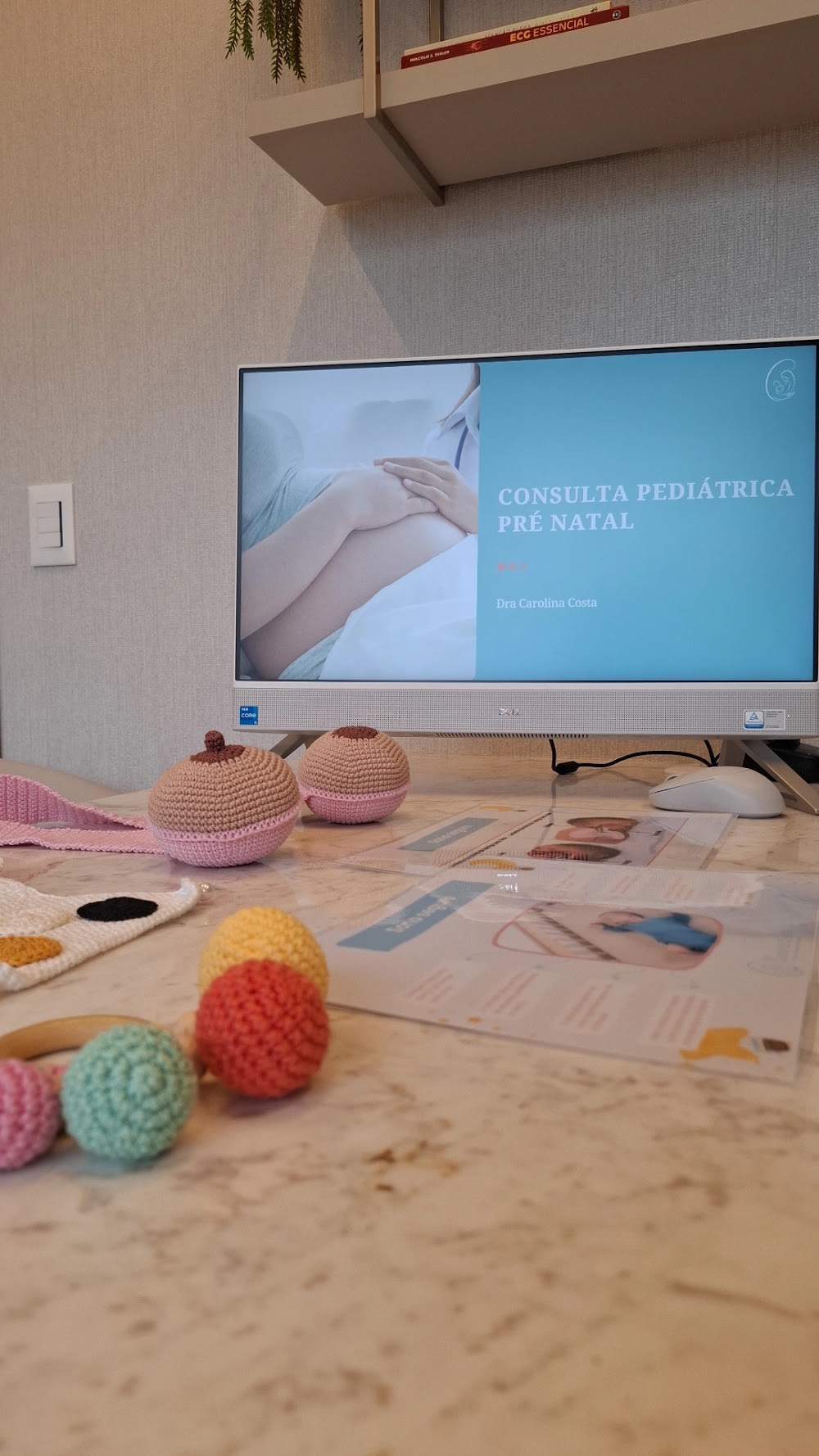 Mesa do consultório com o monitor exibindo material da consulta pediátrica pré-natal, ao lado de brinquedos de bebê