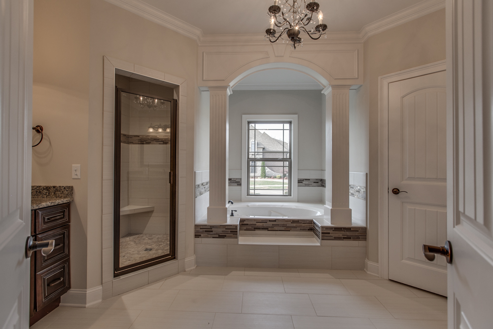 Bathroom interior in a Marymont residential construction project in Middle Tennessee, showing a built-in bathtub under an arch, a glass shower, and tiled flooring.