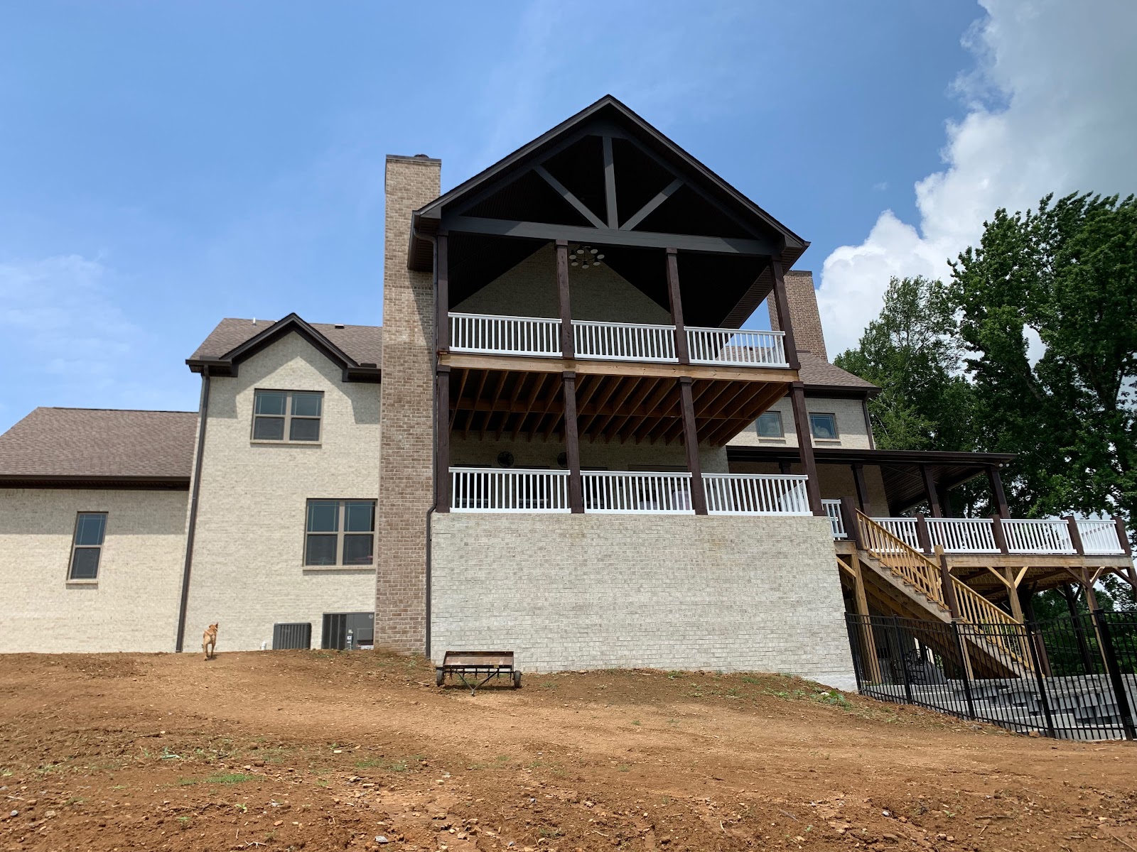 Rear exterior of a light brick house featuring a multi-level deck with white railings and dark support beams above a dirt slope.