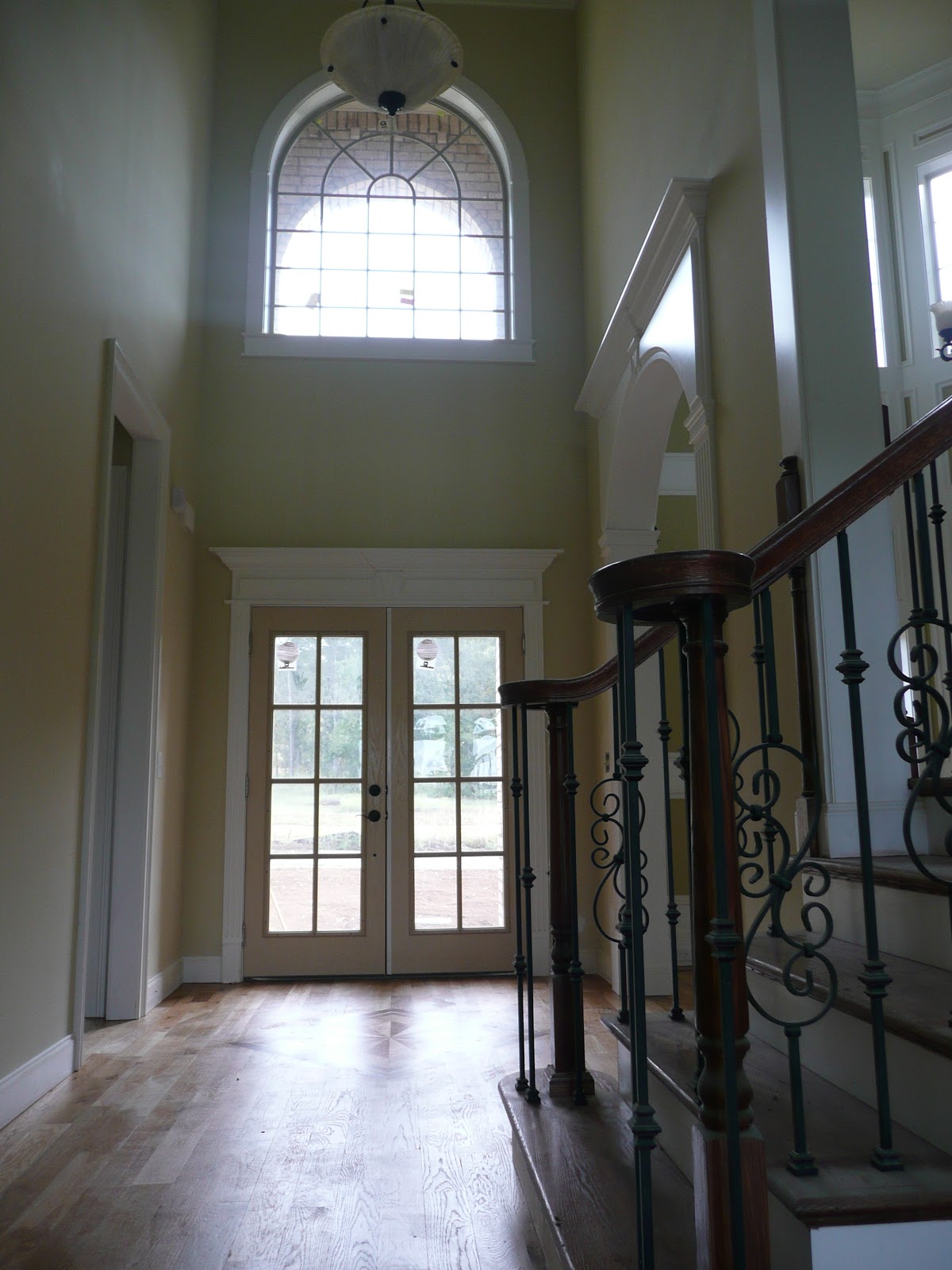 Interior entryway featuring double glass doors, a large arched window above, hardwood floors, and a wooden staircase with decorative metal balusters.