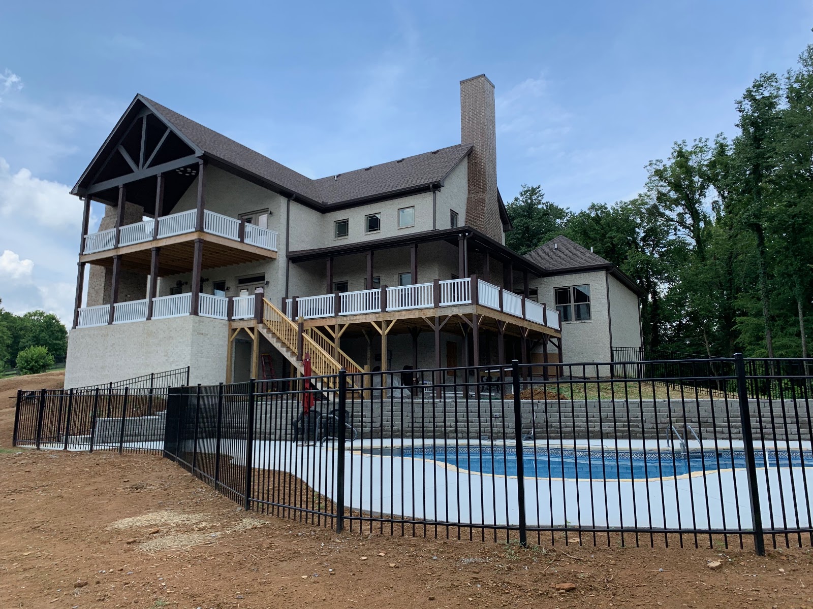 Rear exterior of a light brick house with multi-level decks and white railings, overlooking a fenced-in swimming pool.