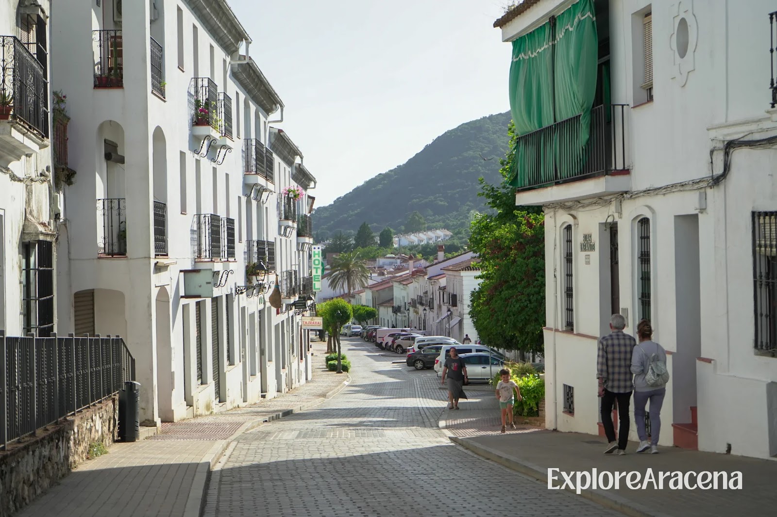 Pedestrians walking along a street of whitewashed buildings in Aracena, with cars parked on both sides.