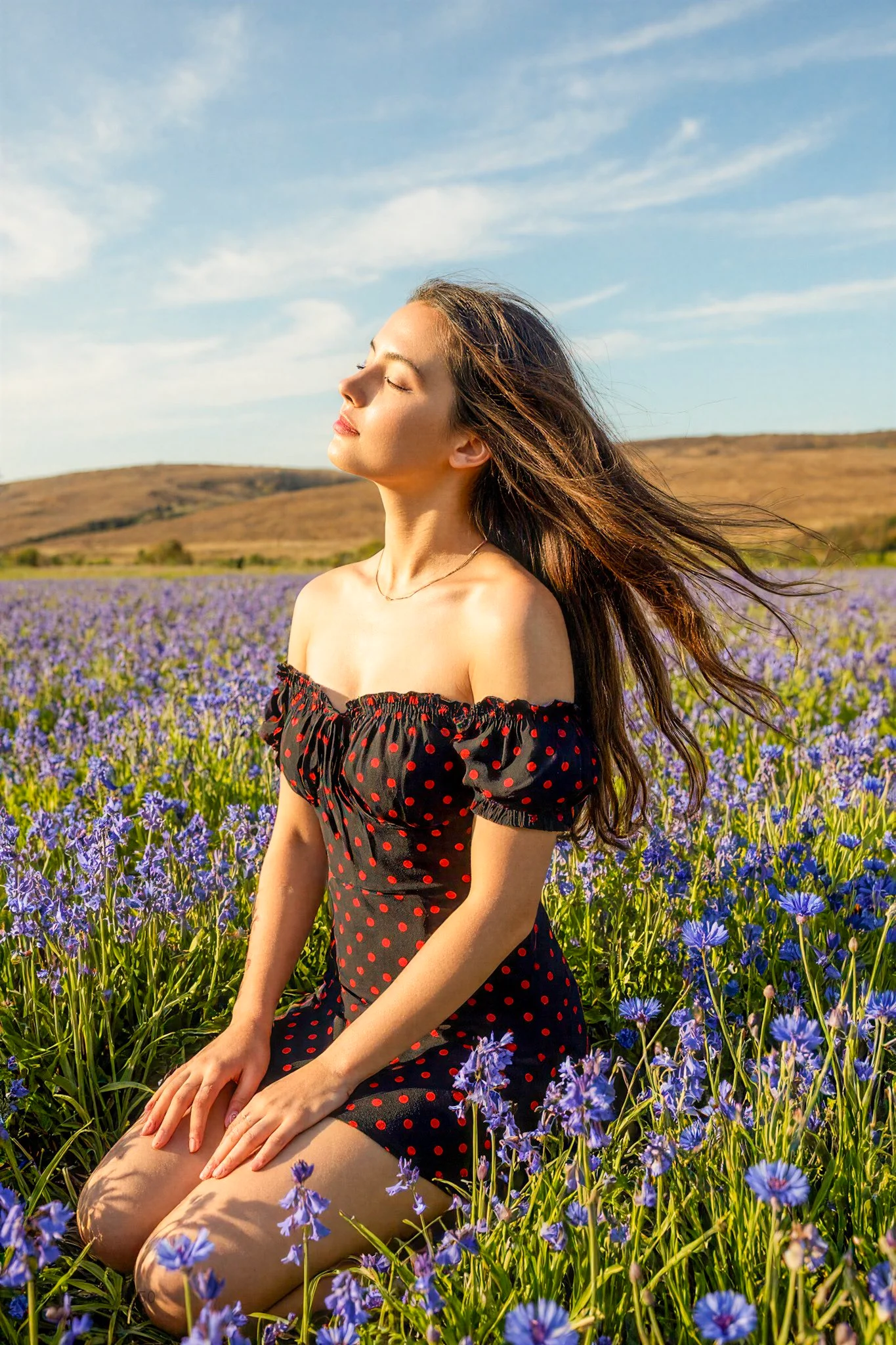 Plano medio completo (de rodillas) de una joven con el pelo largo y alborotado por el viento en un campo de flores silvestres.
