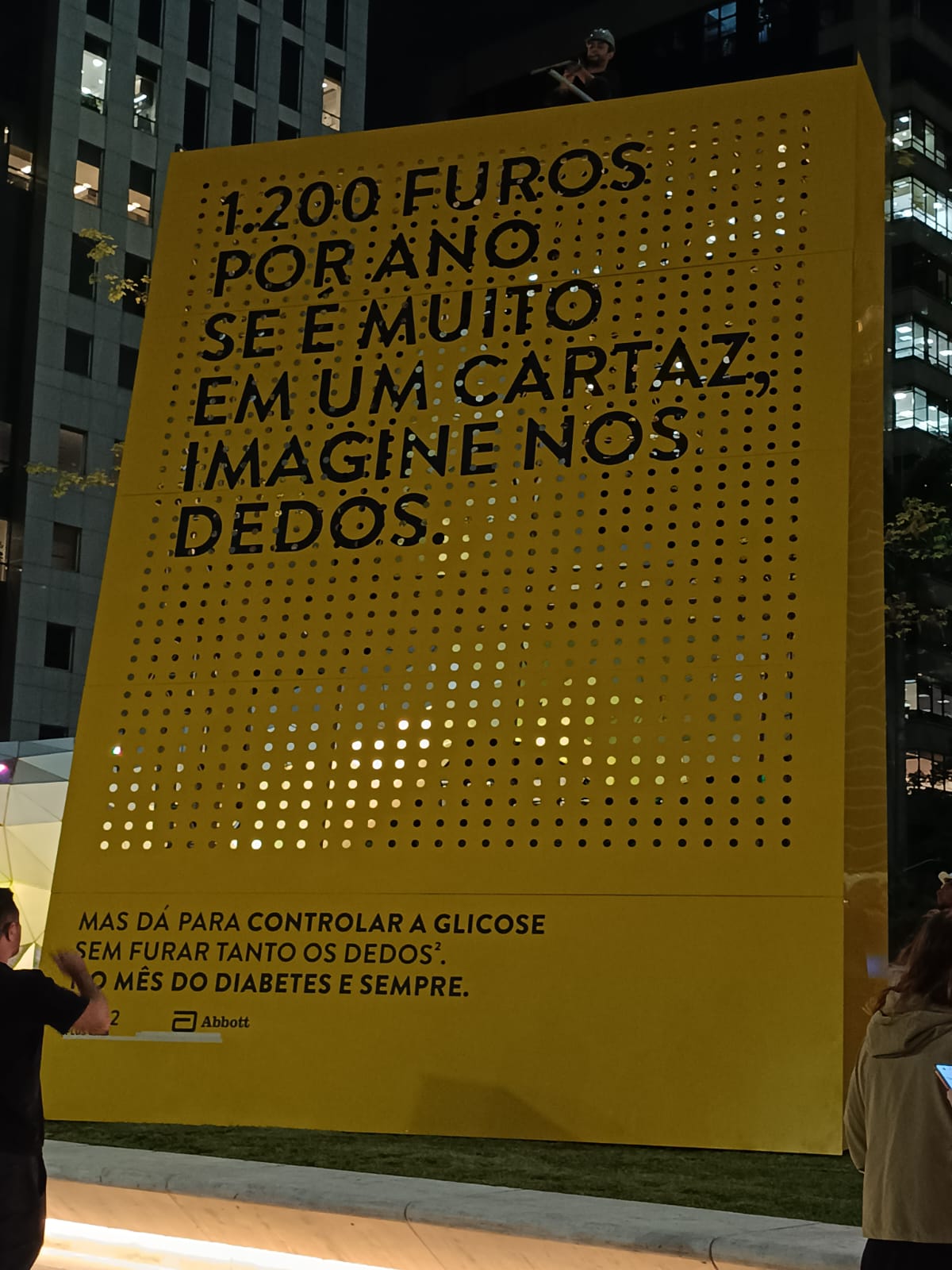 Close-up do painel amarelo da Abbott com texto '1.200 furos por ano' e perfurações, iluminado à noite.