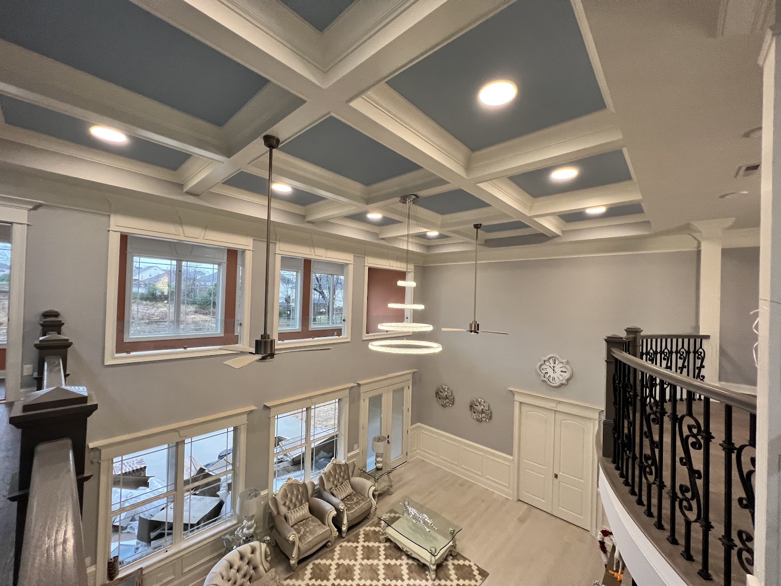 High-angle view from a balcony overlooking a residential living area with a blue-paneled coffered ceiling, large windows, and a tiered ring chandelier.