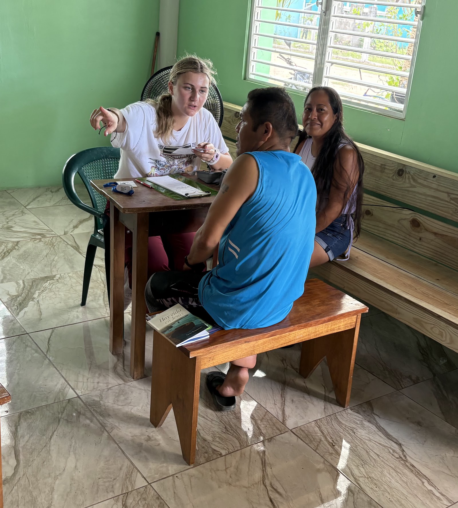 A team member talking with a man and a woman across a table at the Belize clinic; a Spanish New Testament sits on the bench