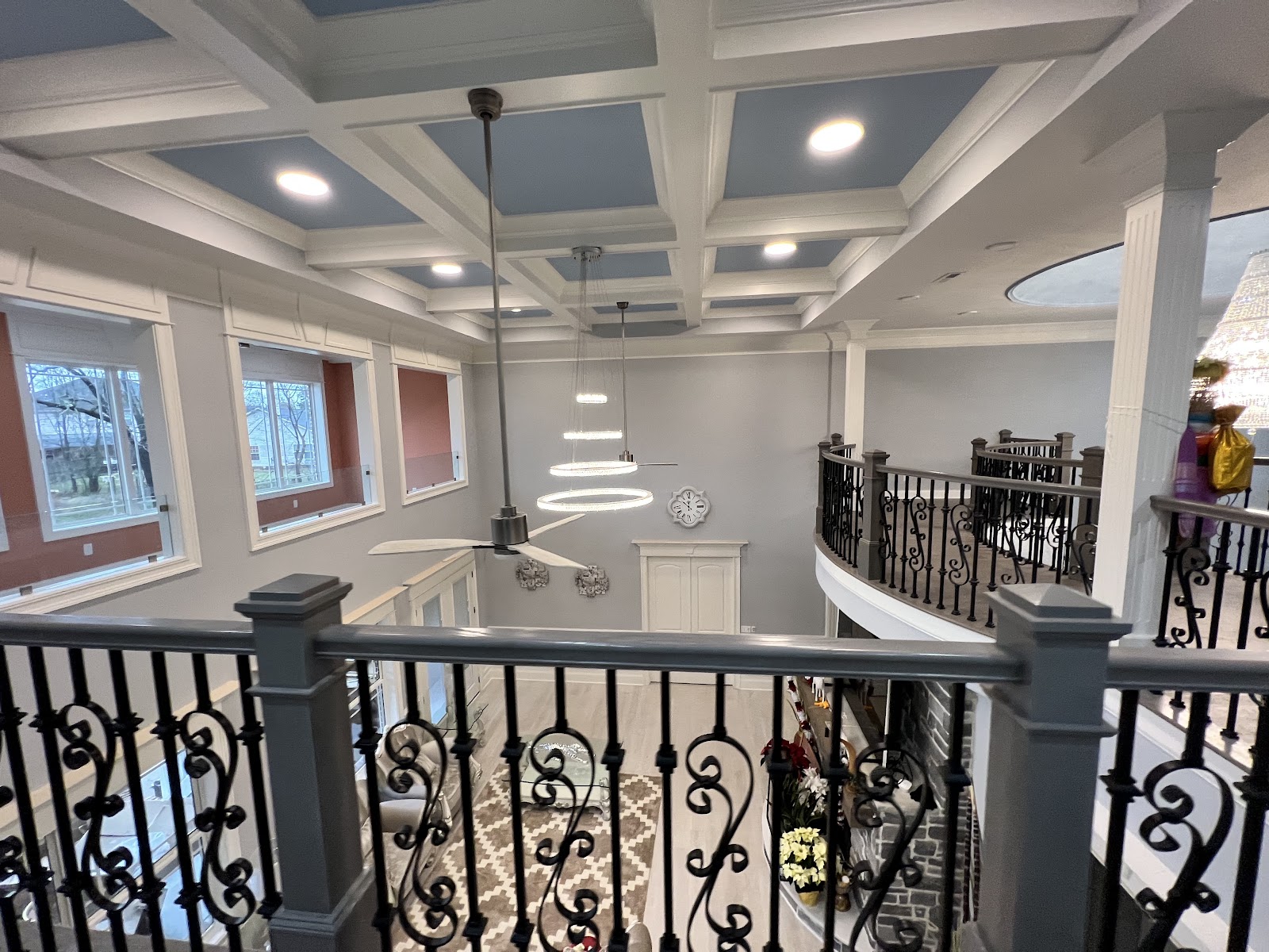 View from an upper-level balcony with wrought iron railing looking down into a living space. The ceiling features a white coffered design with blue-painted panels and recessed lights.