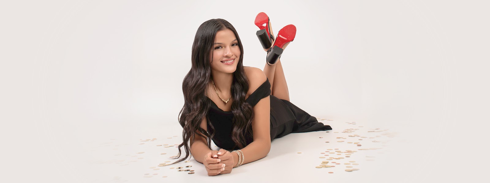 Smiling woman in a black dress lying on a white studio floor with red-soled heels up, surrounded by scattered confetti.