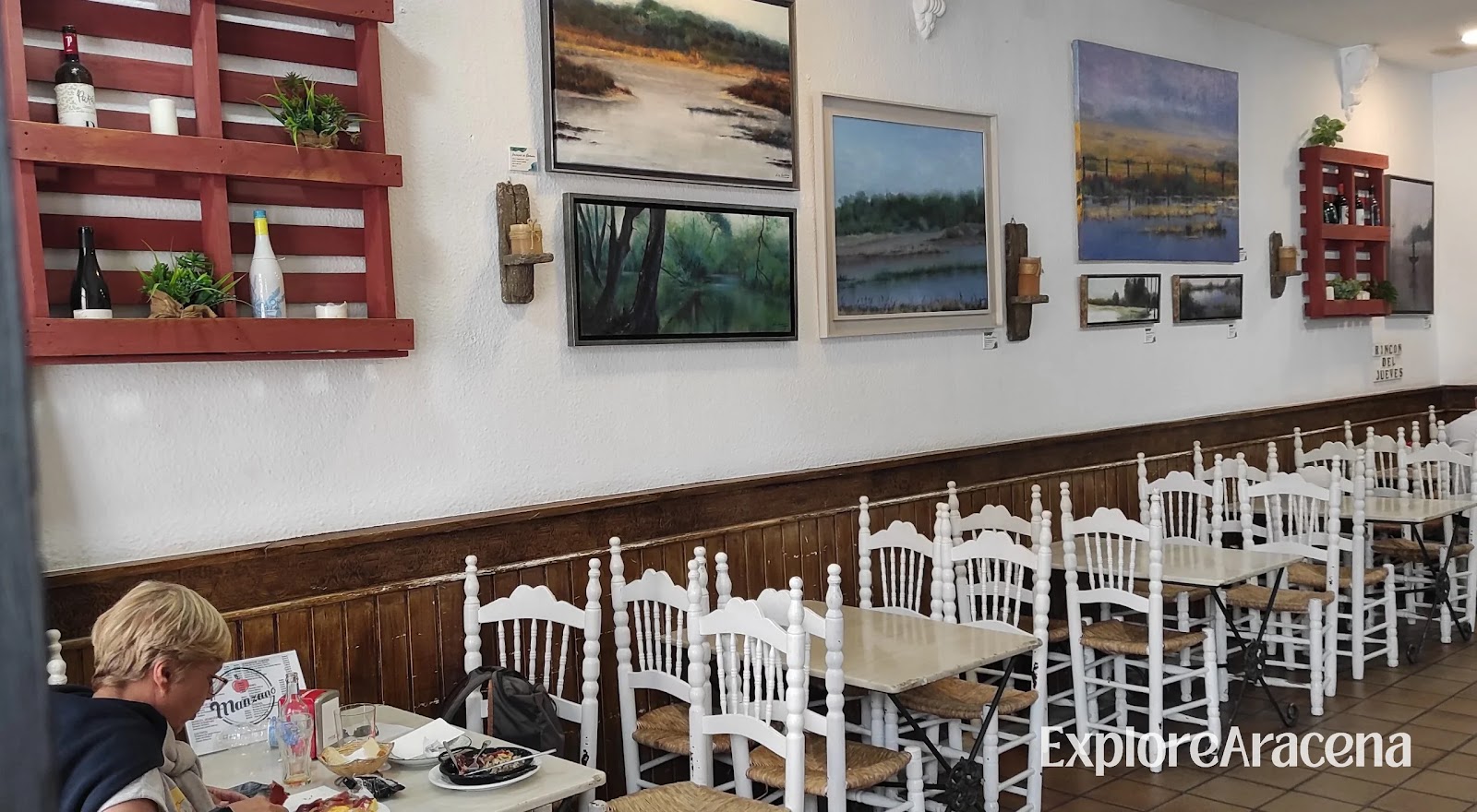 Interior of Café-Bar Manzano in Aracena, showing rustic white chairs, wood panelling, and a wall of local landscape paintings