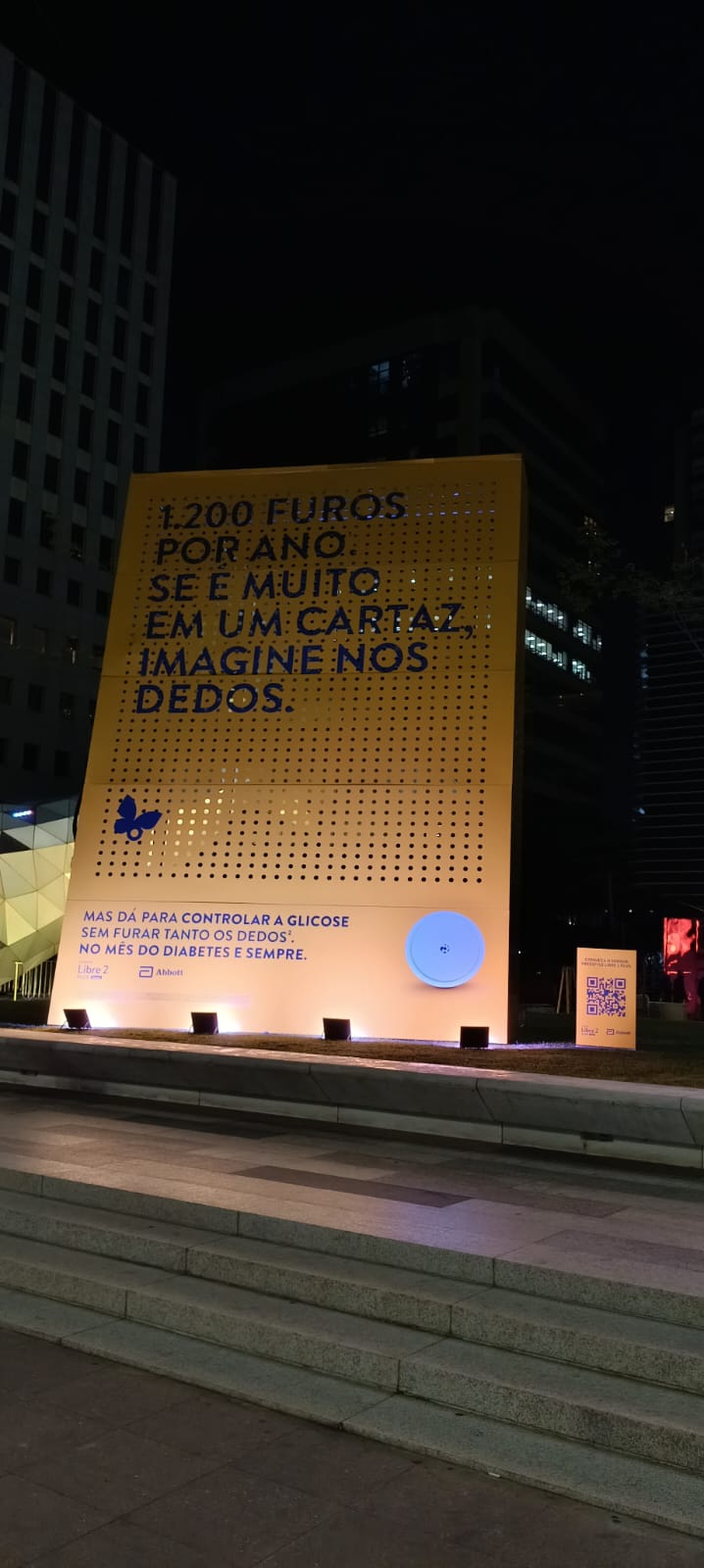 Visão ampla do painel da Abbott em praça pública, com iluminação cênica na base e totem de QR Code ao lado.