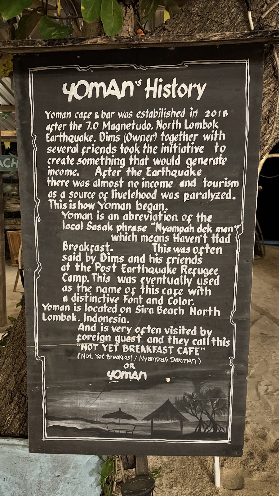 The Yoman's History sign on Sira Beach, North Lombok, explaining the cafe's origin after the 2018 earthquake