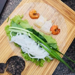 Trải bánh tráng, dùng rau xếp vào, cho thêm bún và các loại rau sống, thêm 2 con tôm và 2 miếng thịt ở góc cuối. Dùng tay cuốn chặt, tiếp theo cho hẹ vào là xong. Lần lượt làm cho hết phần thịt và tôm.