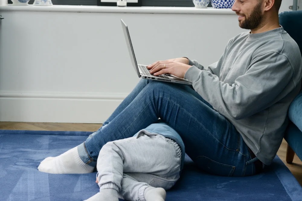 Dad on a laptop beside his toddler, both on a cushioned Momomi mat over a hard floor