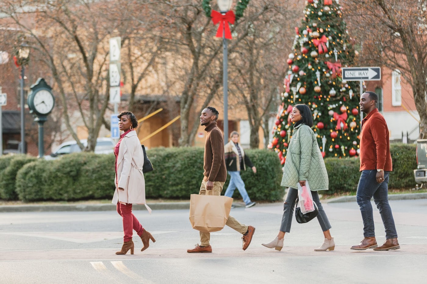 Friends carrying shopping bags cross the street in downtown Ellijay, Georgia in front of a decorated Christmas tree, red bows on the lamp posts, and the town clock