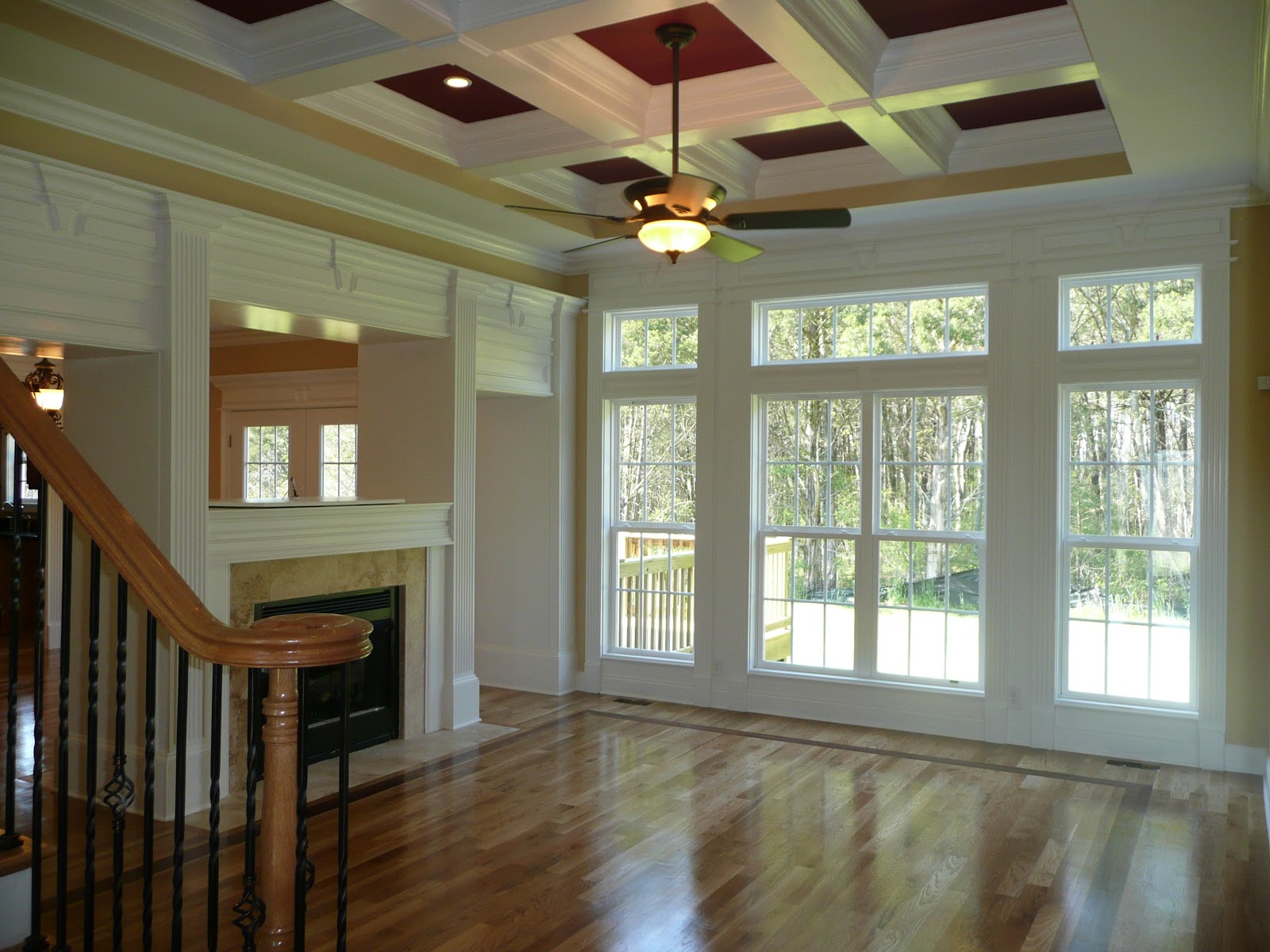 Living area with hardwood floors, a coffered ceiling with red accents, a ceiling fan, and large windows overlooking trees.