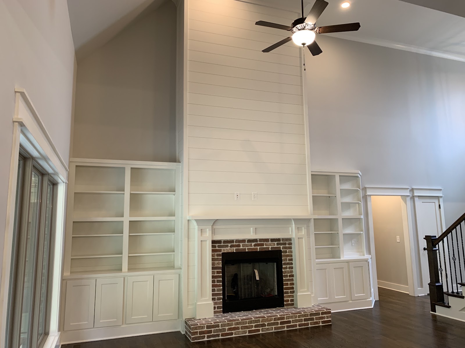Living area with high ceilings, a central brick fireplace with a white mantel and shiplap wall, flanked by built-in white bookshelves and cabinets on dark wood floors.