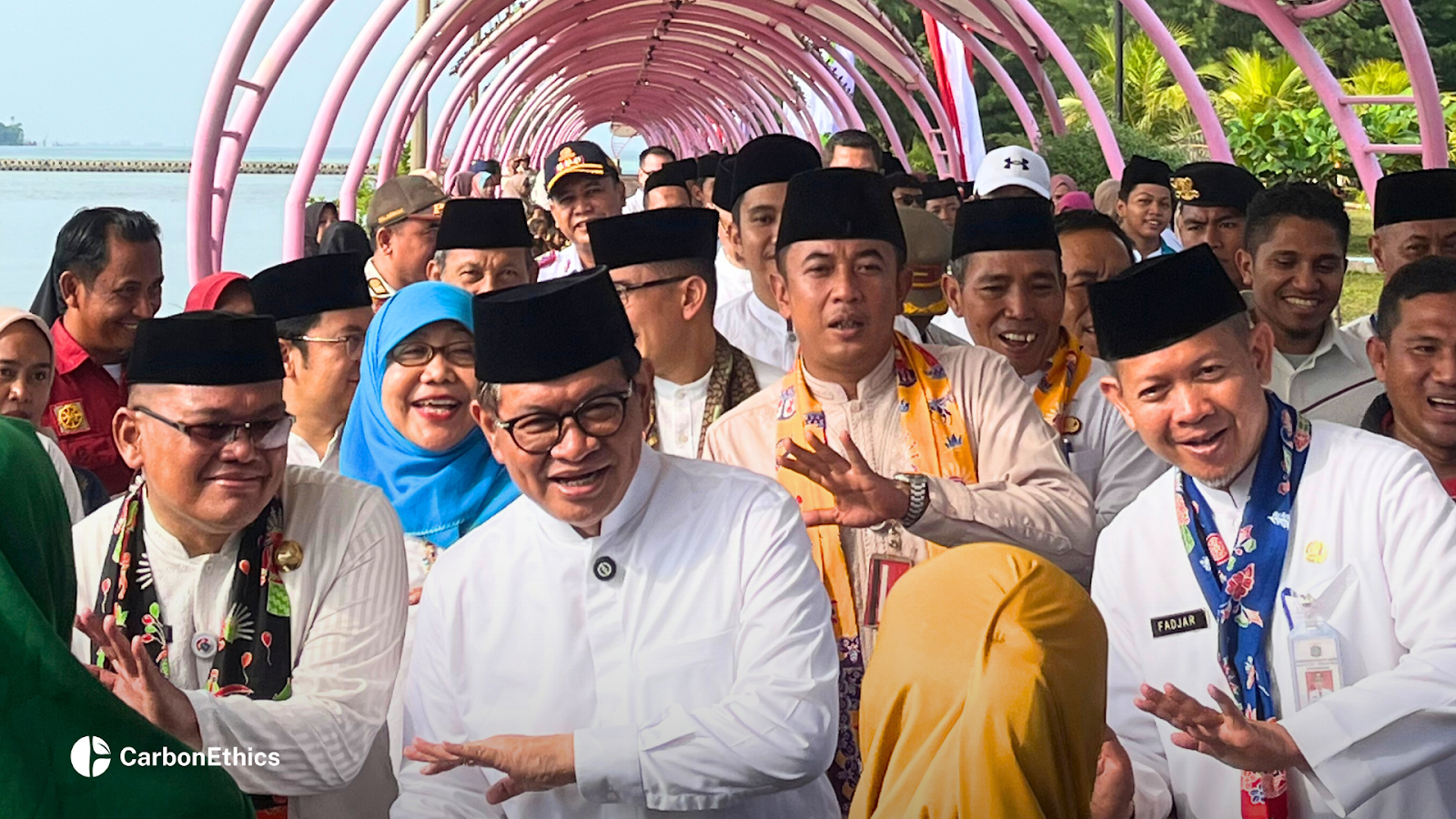 Jakarta Governor Pramono Anung smiling while walking under a pink archway with other officials and local community members during a site visit in Kepulauan Seribu. The atmosphere is celebratory and welcoming.