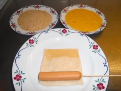 Cuốn như hướng dẫn ở hình dưới. Cho bánh mì trên dĩa, đặt xúc xích từ ngoài cuốn vào, làm lần lượt với xúc xích còn lại. Nhúng bánh mì cuộn xúc xích qua lớp trứng, cuối cùng nhúng qua lớp bột chiên xù.
