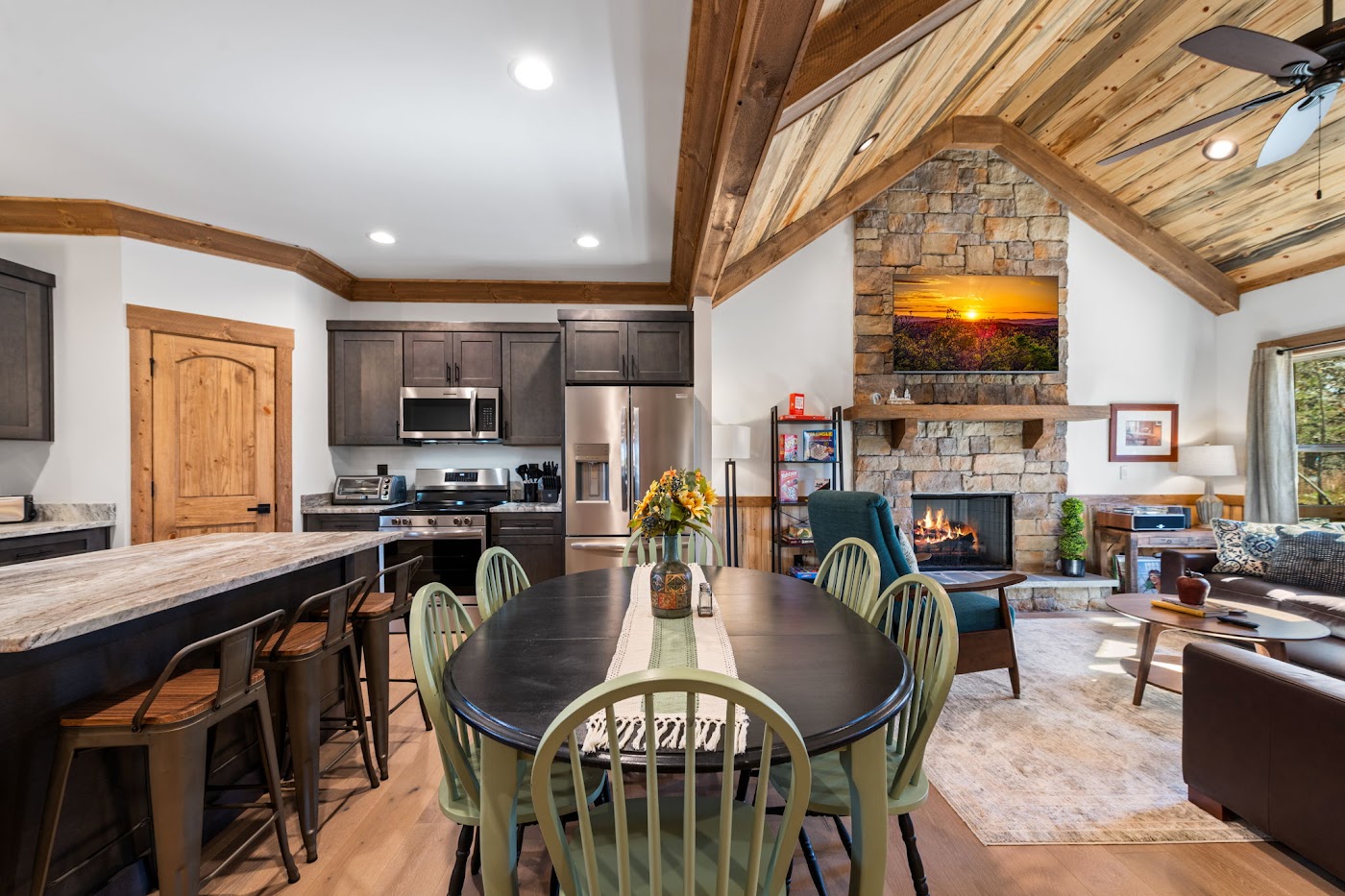 Open kitchen, dining table with green chairs, and stone gas fireplace under a wood-plank vaulted ceiling at Orchard & Vine Mountain Haus in Ellijay, Georgia