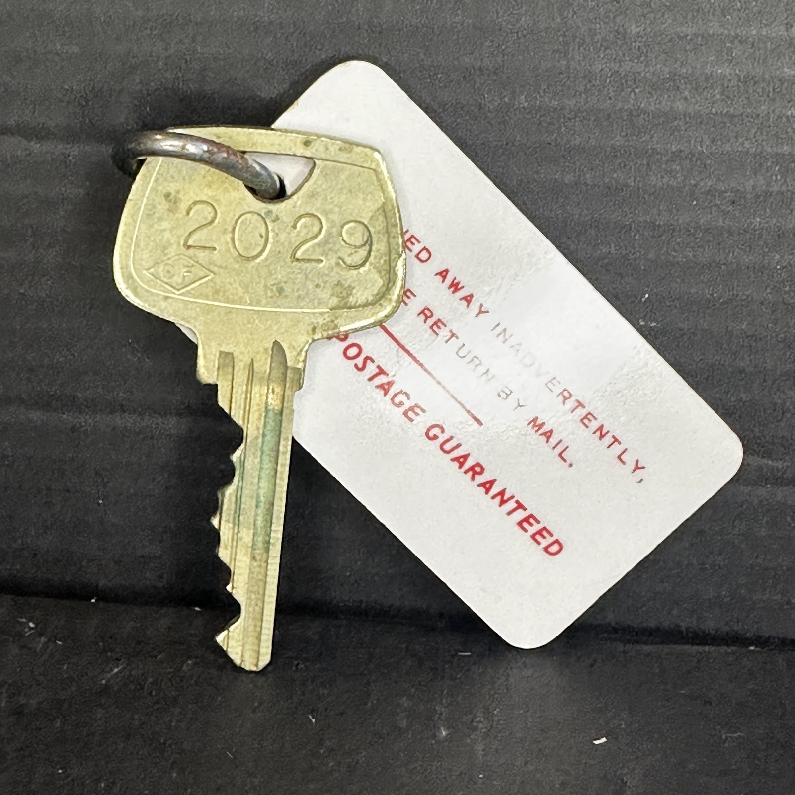Brass Disney "DF" locker key number 2029 on steel ring with original white cardstock return-by-mail tag, showing verdigris patina and age yellowing