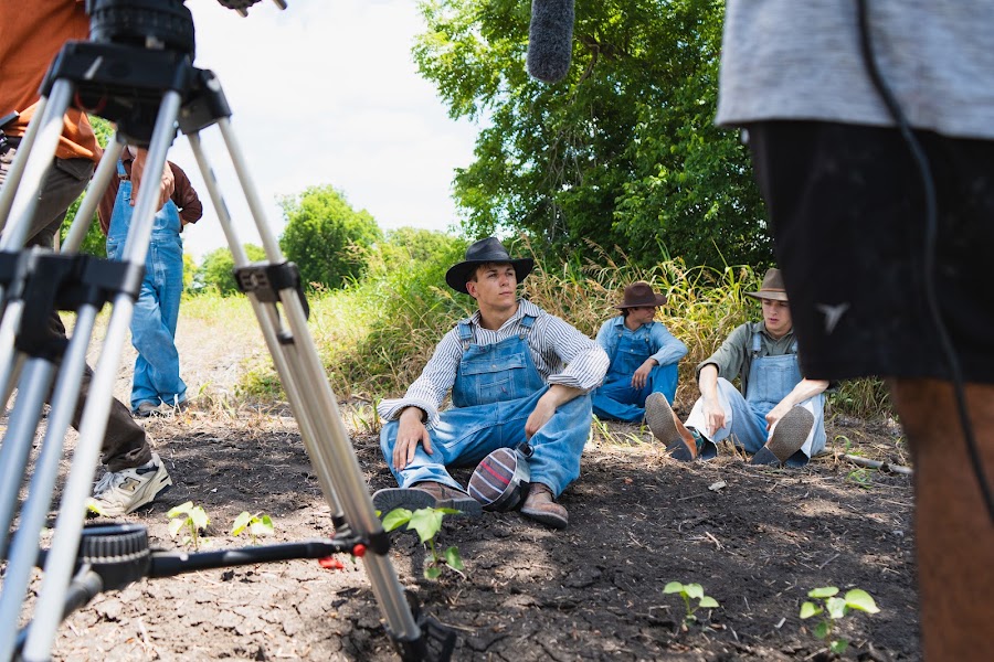Three actors in overalls rest in the dirt beside the camera tripod