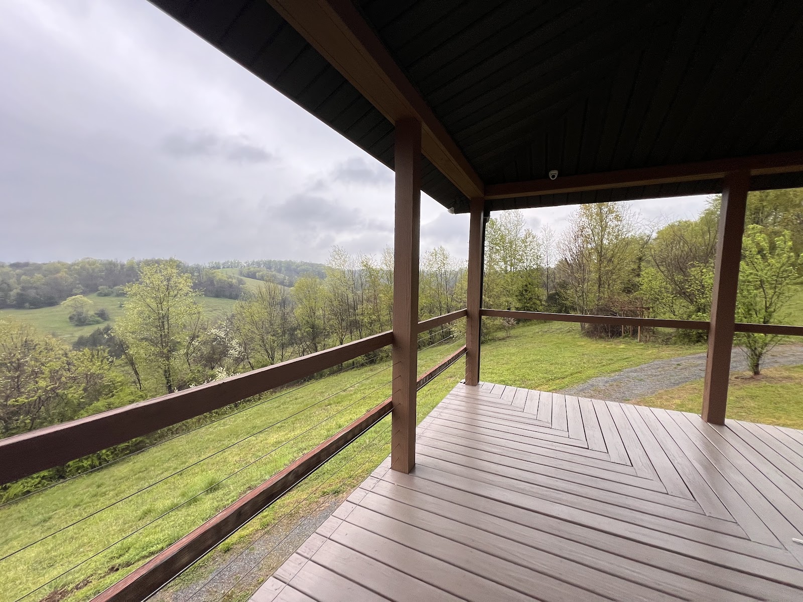Wooden deck with chevron-patterned floorboards and a cable railing overlooking a green, hilly rural landscape under a cloudy sky.