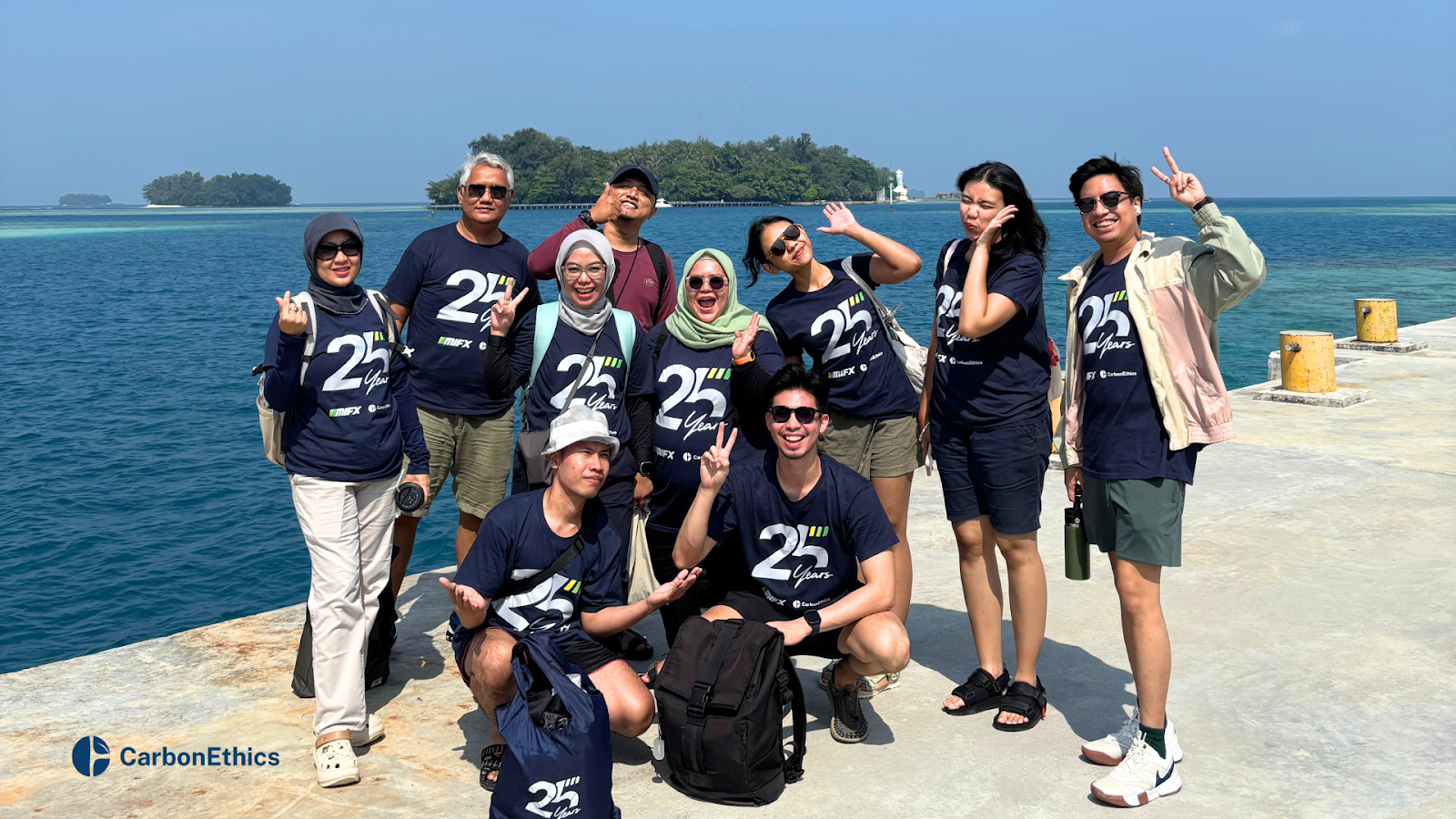 MIFX team posing cheerfully on a dock with the sea and islands behind them, wearing “25 Years” anniversary shirts for their collaboration with CarbonEthics.
