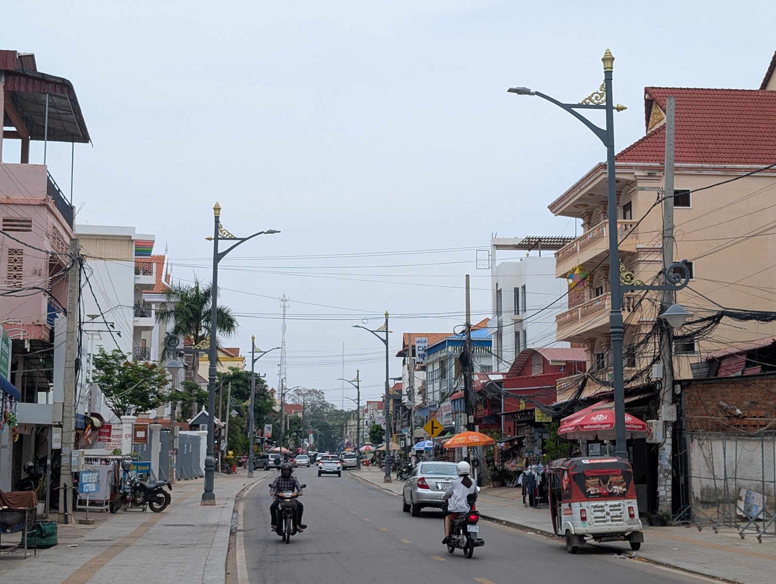 A street in Siem Reap with four story apartment building on
                 either side with balconies overlooking motorbikes, tuk tuks,
                 a few cars, and umbrellas below. Utility poles and tall light poles
                 sprout from the side walk, and cables zig zag between them
                 above the road. A tall radio tower is seen in the background.
                 