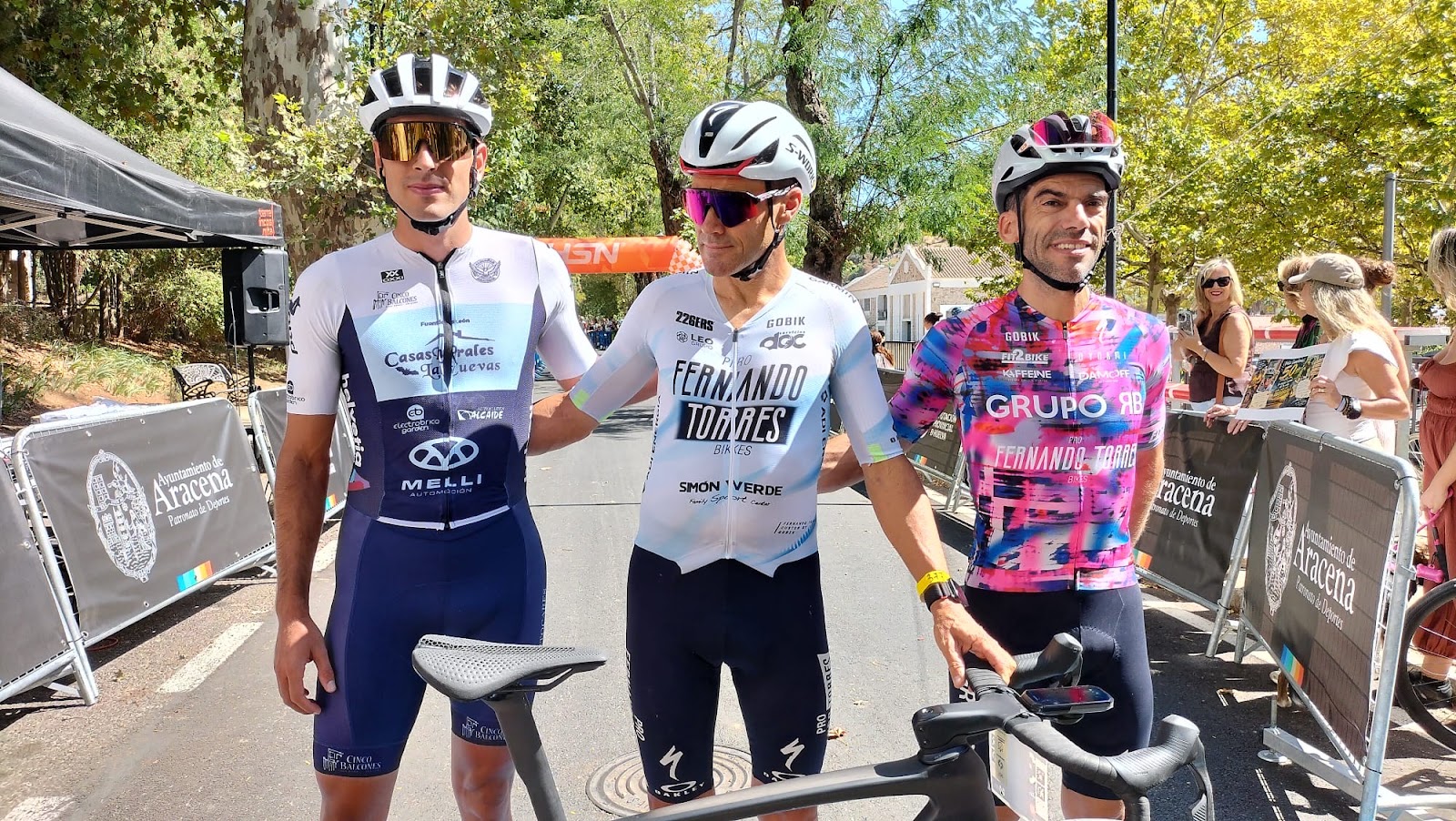 Three cyclists standing with their road bikes and smiling in a Sierra de Aracena village during the HUEX Gran Fondo 2026.