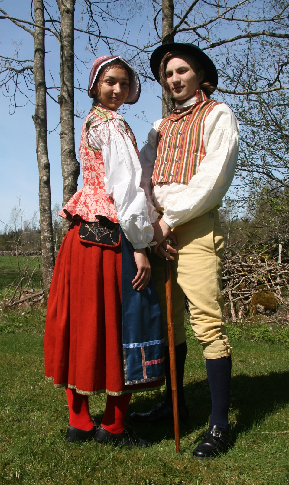 Småland, Västbo. Isabell bär livstycke i damast med mössa i samma tyg. Förkläde i raskimitation med påsydda band. Axel i ylleväst.