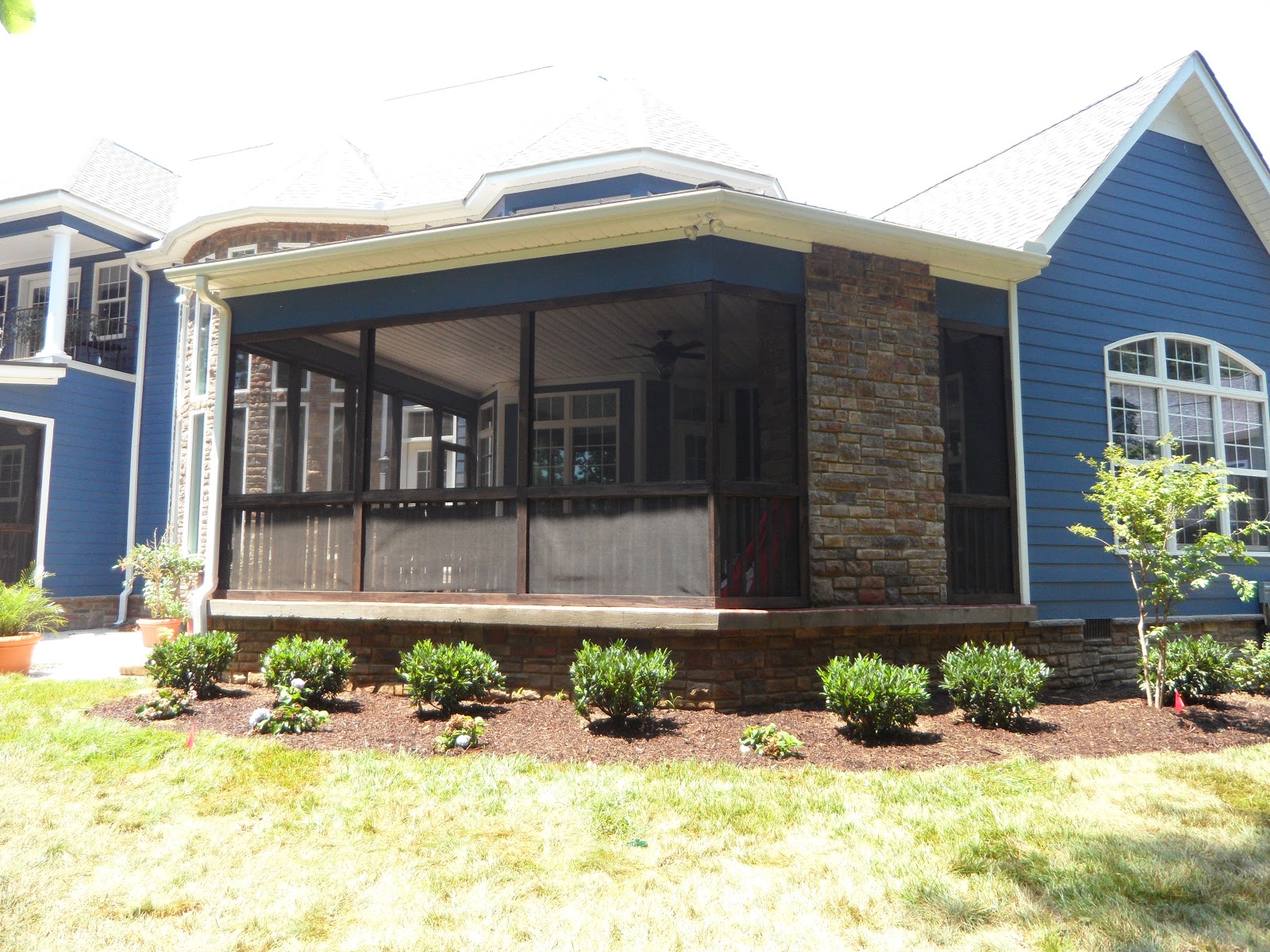 Exterior view of a screened-in porch with stone accents attached to a house with blue siding. Small green bushes are planted in a mulch bed in front.