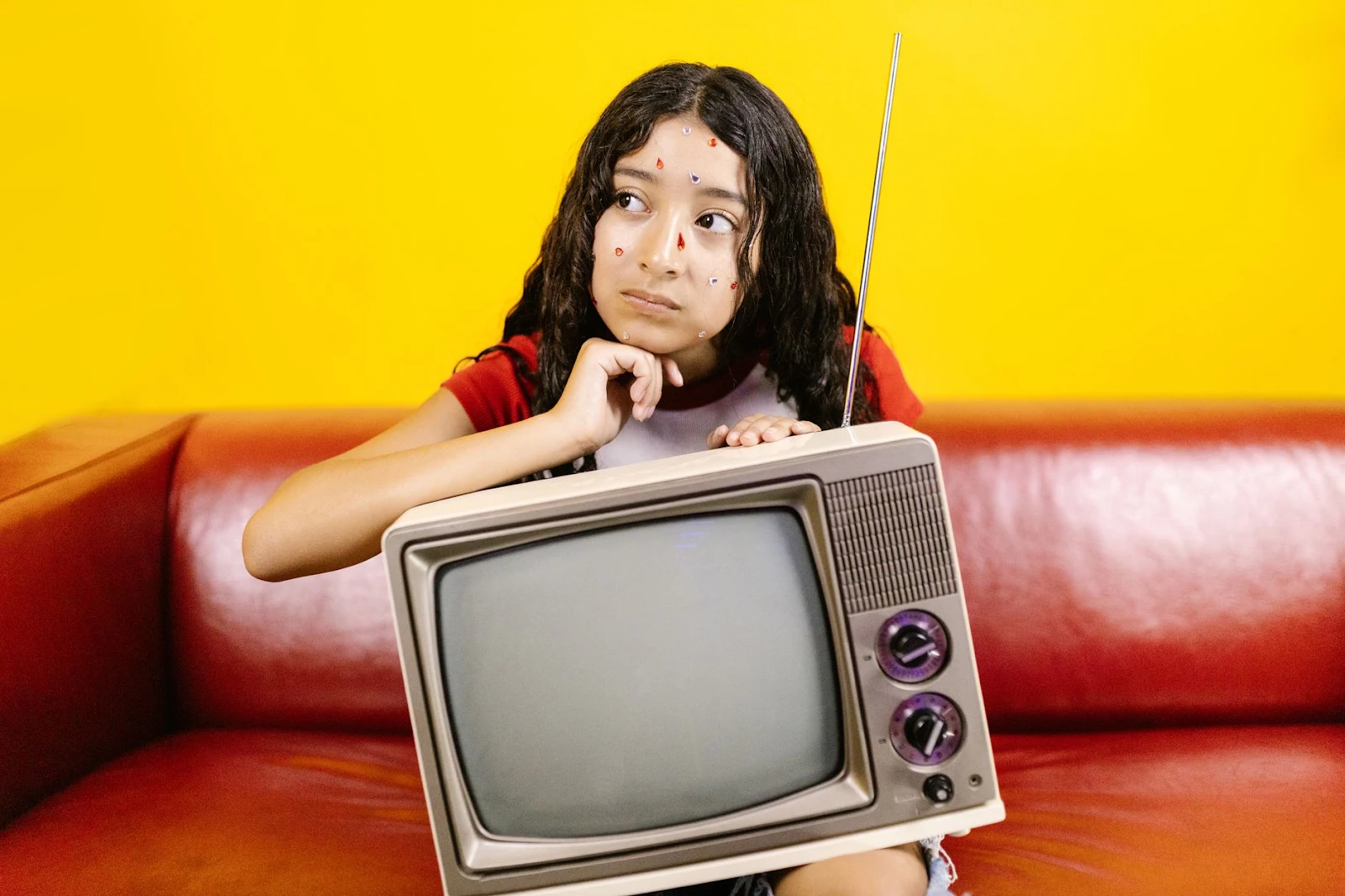 Woman with stickers on face posing with a vintage TV on a red couch with a bright yellow background.