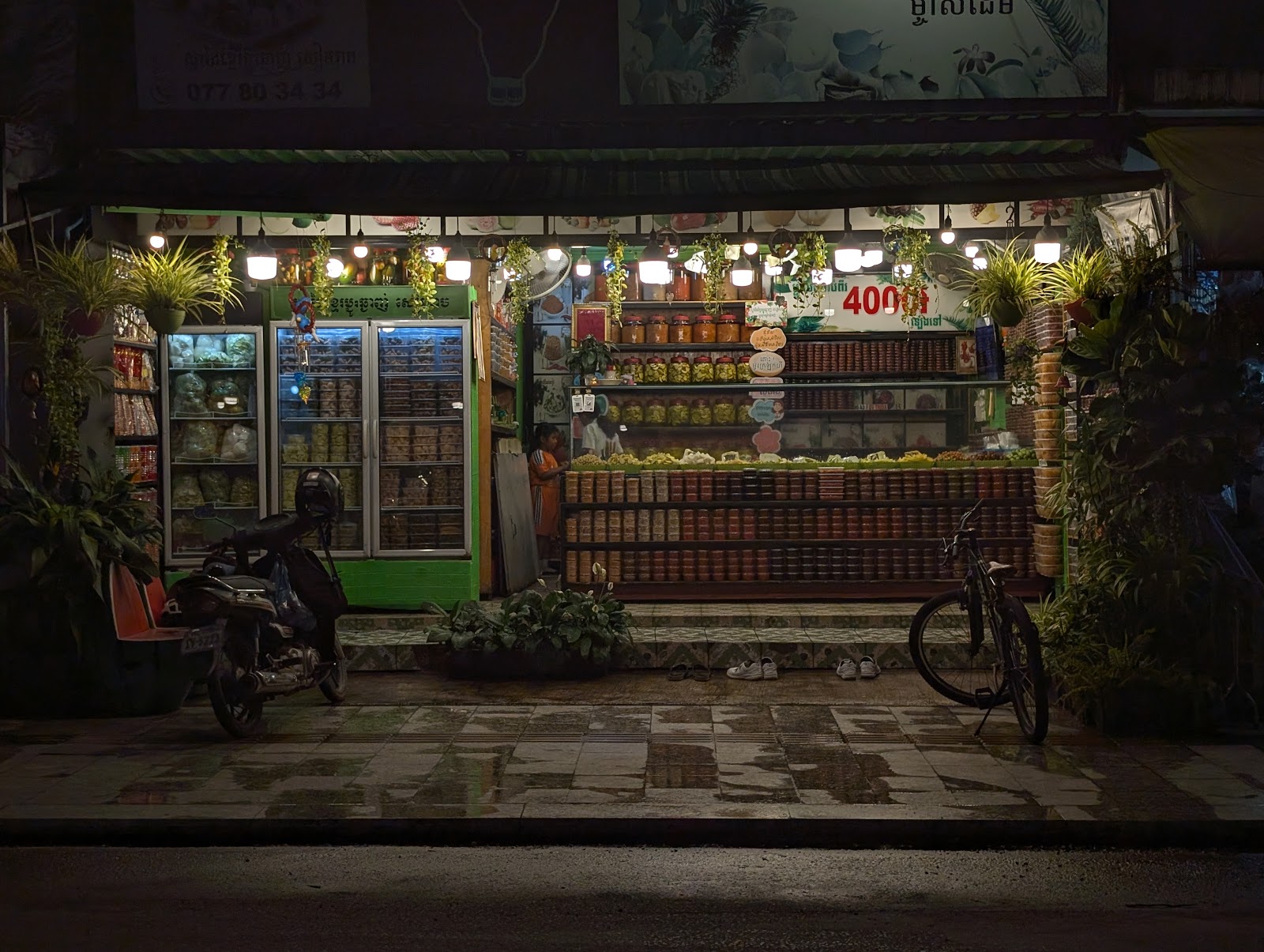 A dim photo showing an open air storefront with shelves and a
                 counter full of a variety of colorful products. Light is from
                 stringlights right under the store's signage. A bike and a
                 motorbike are parked in front.