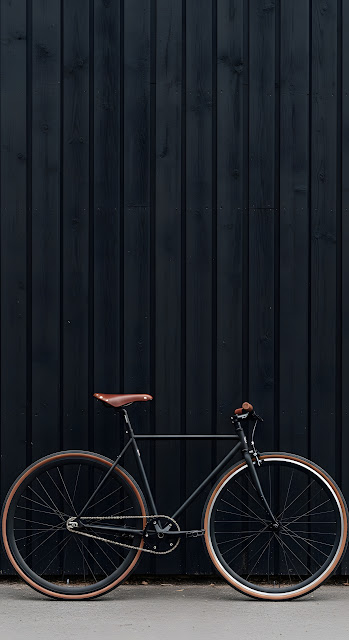 Minimalist Black Single-Speed Bicycle with Brown Tires Against Dark Textured Wall - Free mobile Wallpaper