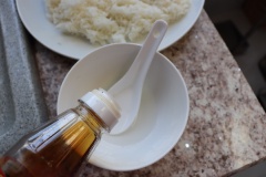 Cơm trắng nóng múc ra dĩa để dễ trộn hơn, bạn cũng có thể bỏ vào tô. Pha nước trộn cơm: cho giấm vào chén cùng 1 xíu muối rồi trộn đều cùng dầu mè, nước tương và đường.