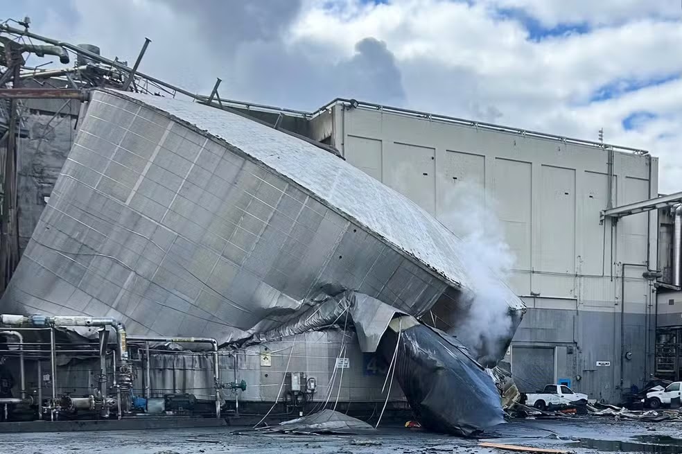Vista aérea de fábrica industrial às margens de rio com fumaça e equipes de emergência