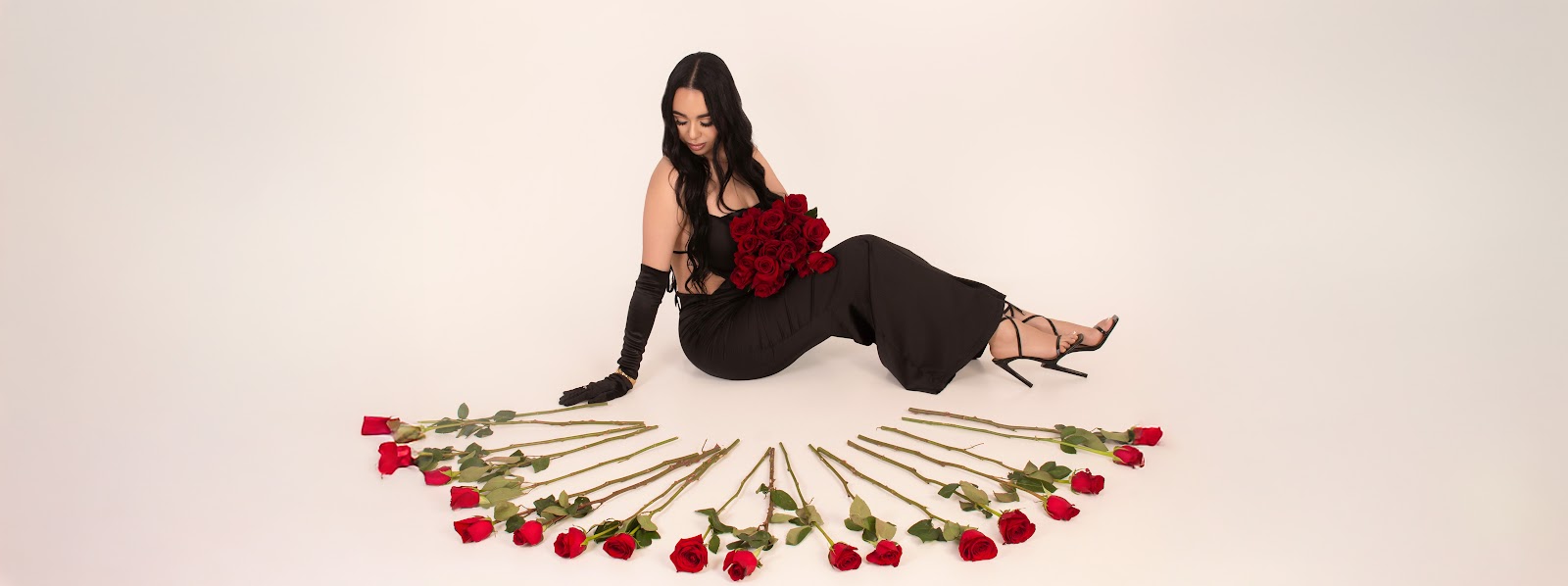 Woman in a black dress and gloves seated on a white studio floor, holding a bouquet of red roses with additional roses arranged in a semicircle in front of her.