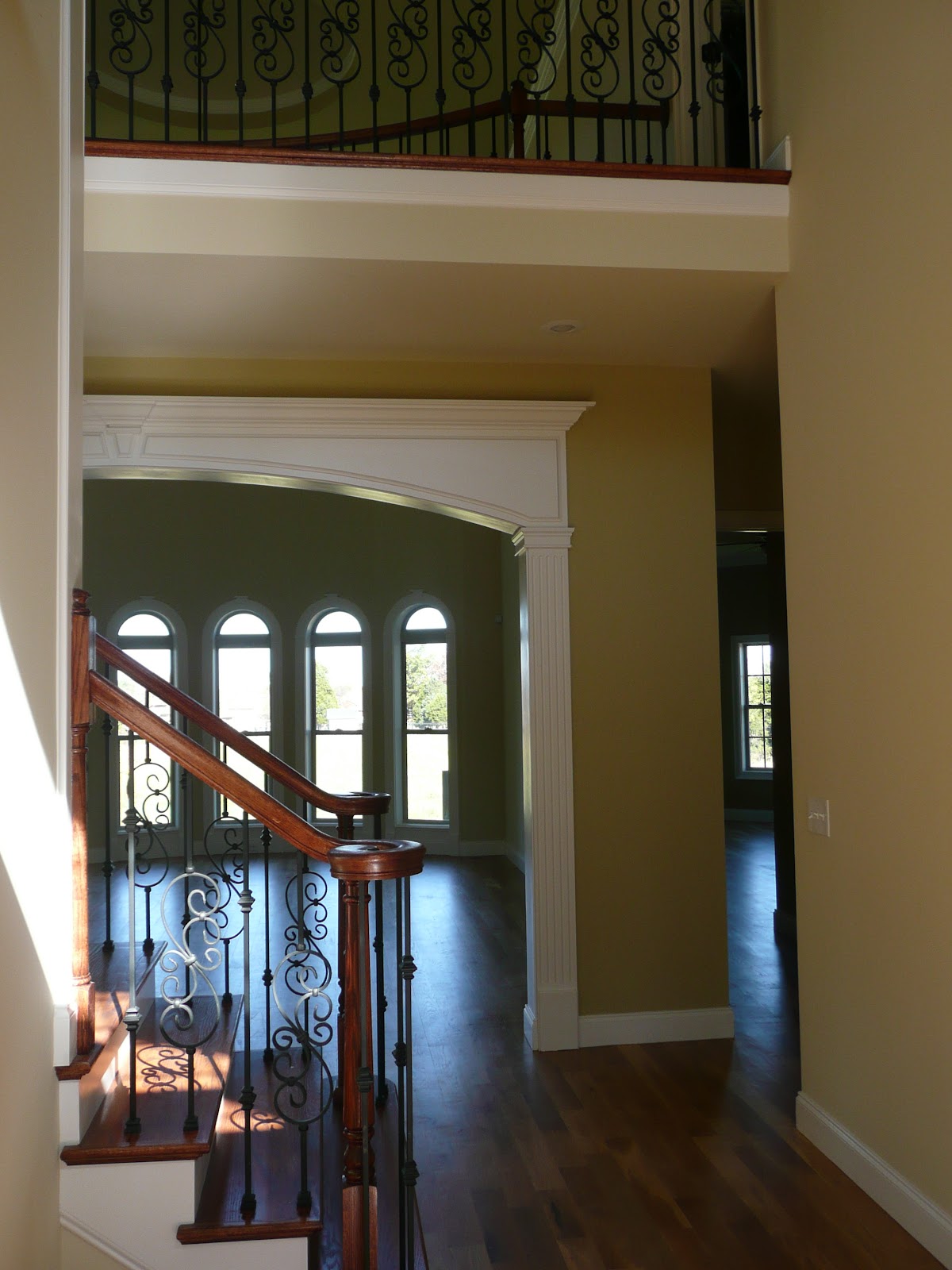 View through a white interior archway toward a room with four arched windows, featuring a staircase with decorative metal balusters in the foreground.