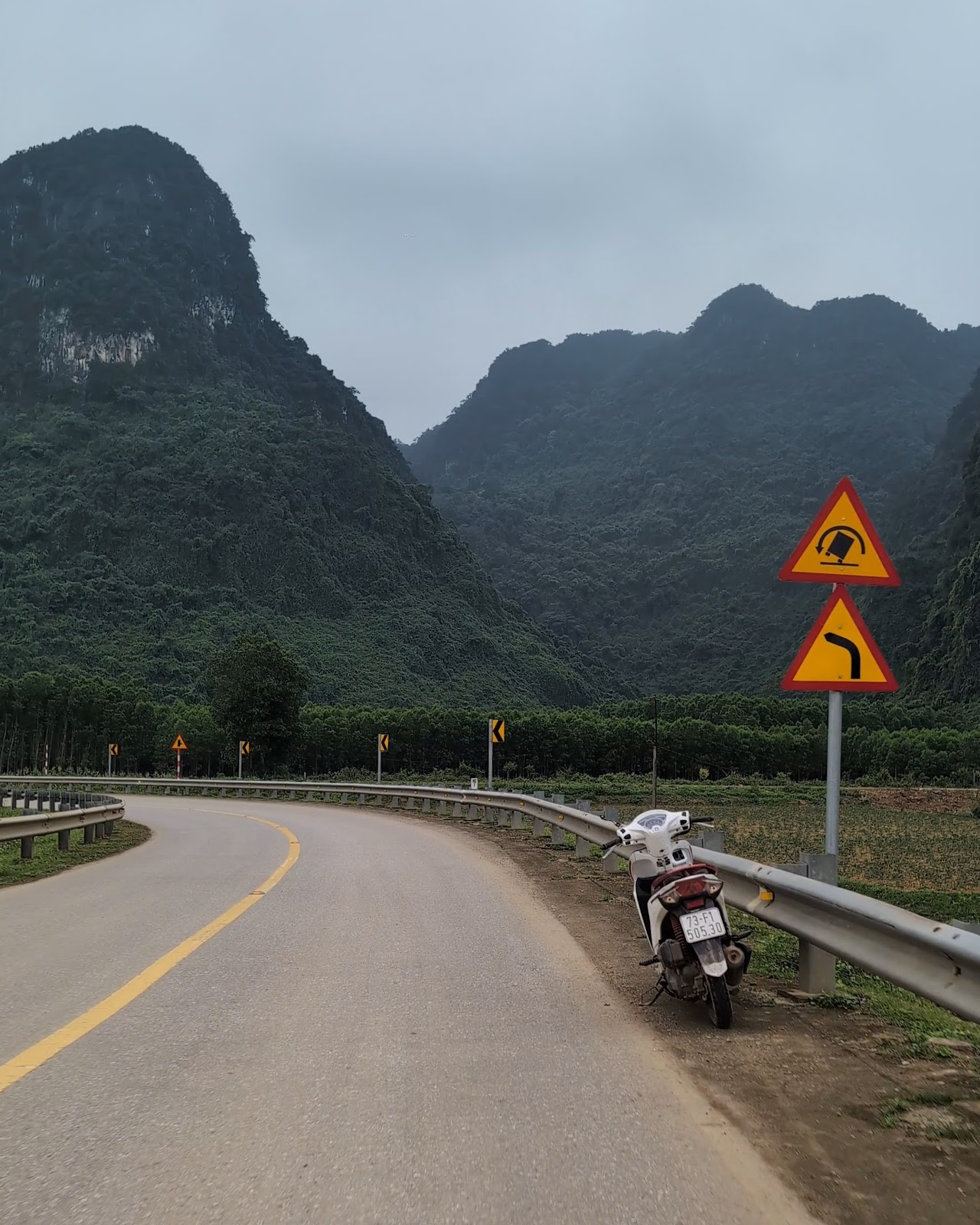 A photo from the road of a curving but empty road with a lone motorbike on
                 the shoulder. In the background is a cloudy sky with some large, lush mountains
                 cresting in the not too far off distance.