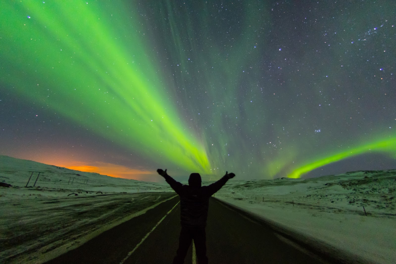 Traveler standing beneath vivid green Northern Lights in a snowy Arctic landscape.