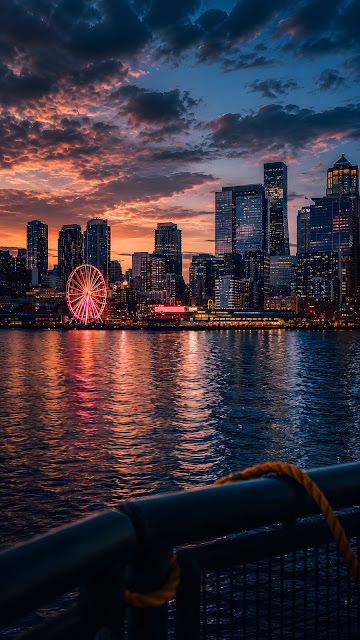City waterfront skyline with brightly lit ferris wheel reflected in water under glowing pink and orange sunset clouds.