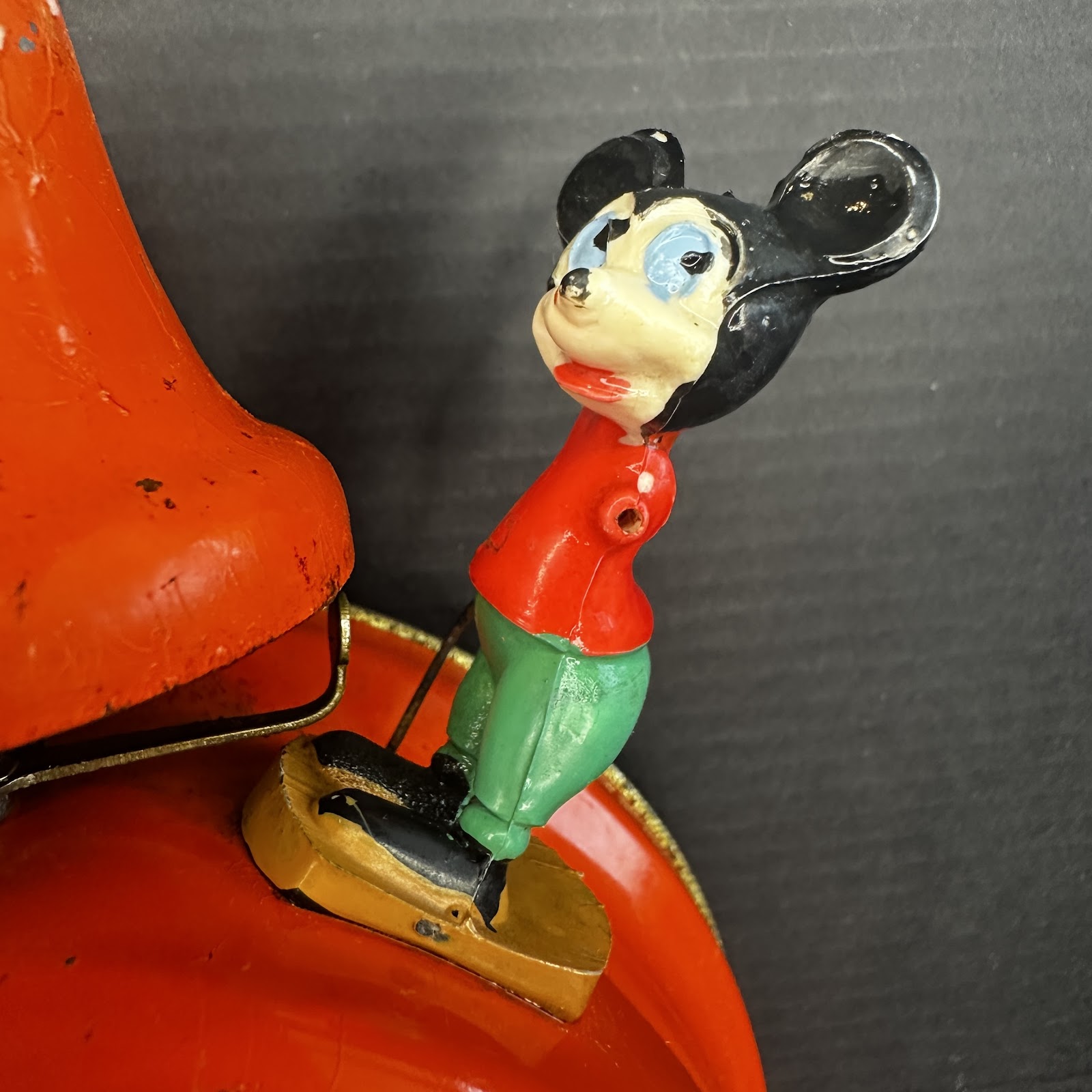 Pre-war celluloid and plastic Pie-Eyed Mickey Mouse figure with red shirt and green pants, mounted on gold metal bracket and red tin base, showing age-appropriate paint wear and facial crazing