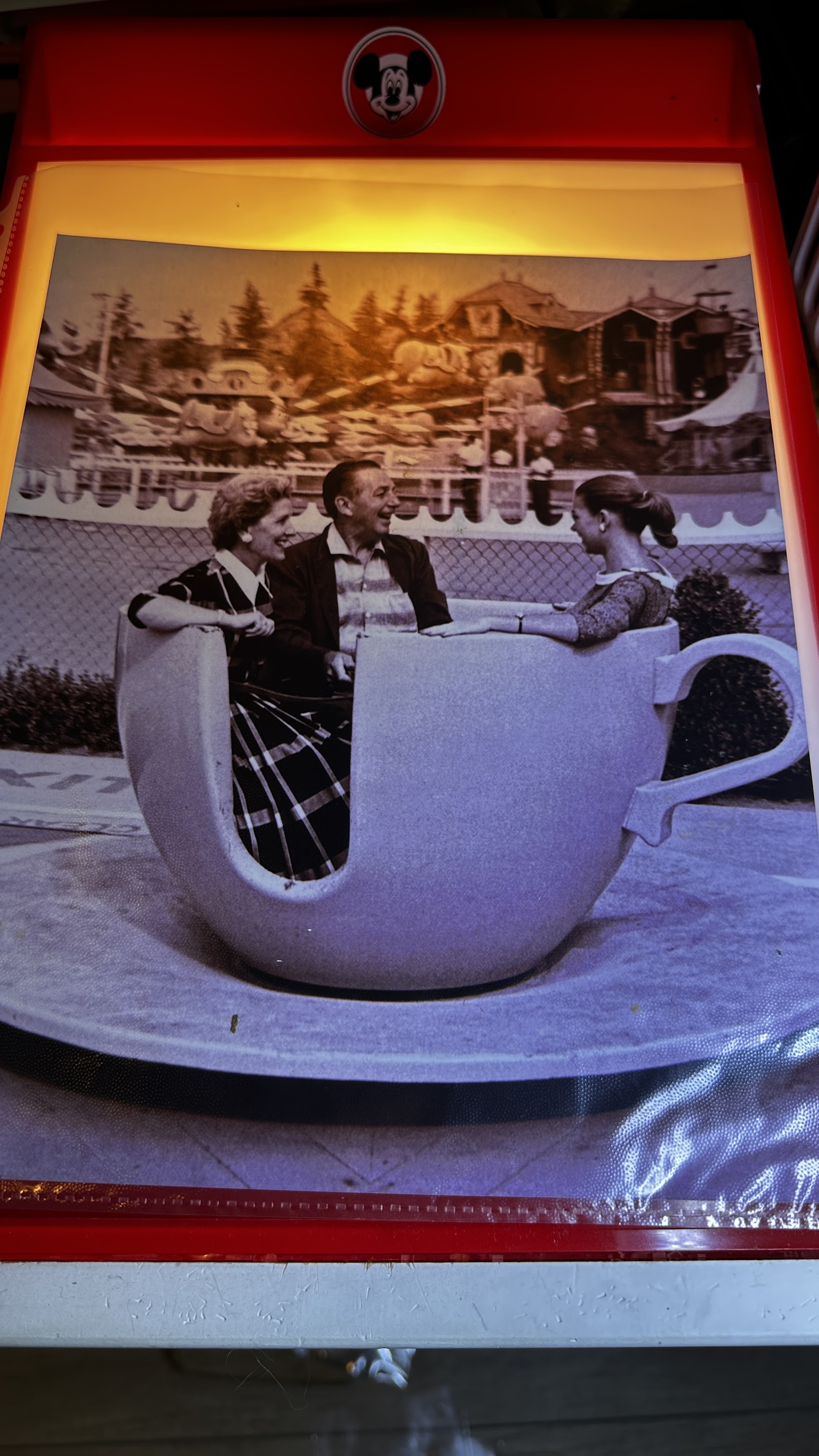 Red plastic light-up photo viewer with pie-eyed Mickey Mouse logo on top, displaying a sepia-toned transparency of Walt Disney, Lillian Disney, and Diane Disney seated in a Mad Tea Party teacup at Disneyland with Fantasyland visible in the background