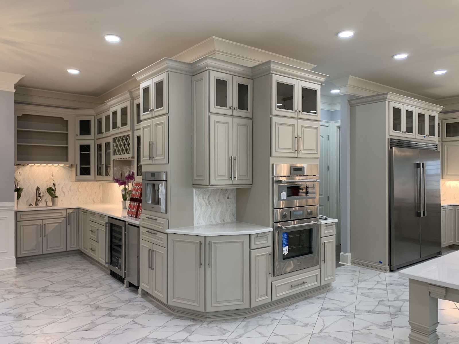Kitchen with light grey cabinetry, white marble-patterned tile flooring, and stainless steel appliances including a double wall oven and a large refrigerator.