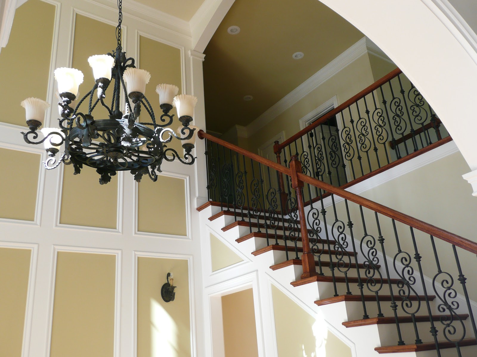 Interior staircase with wooden handrails and wrought iron balusters, featuring an ornate chandelier and decorative wall paneling.