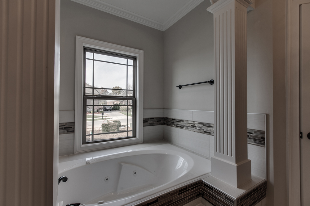 A white jetted bathtub with a decorative tile border, a fluted column, and a window with a black frame.