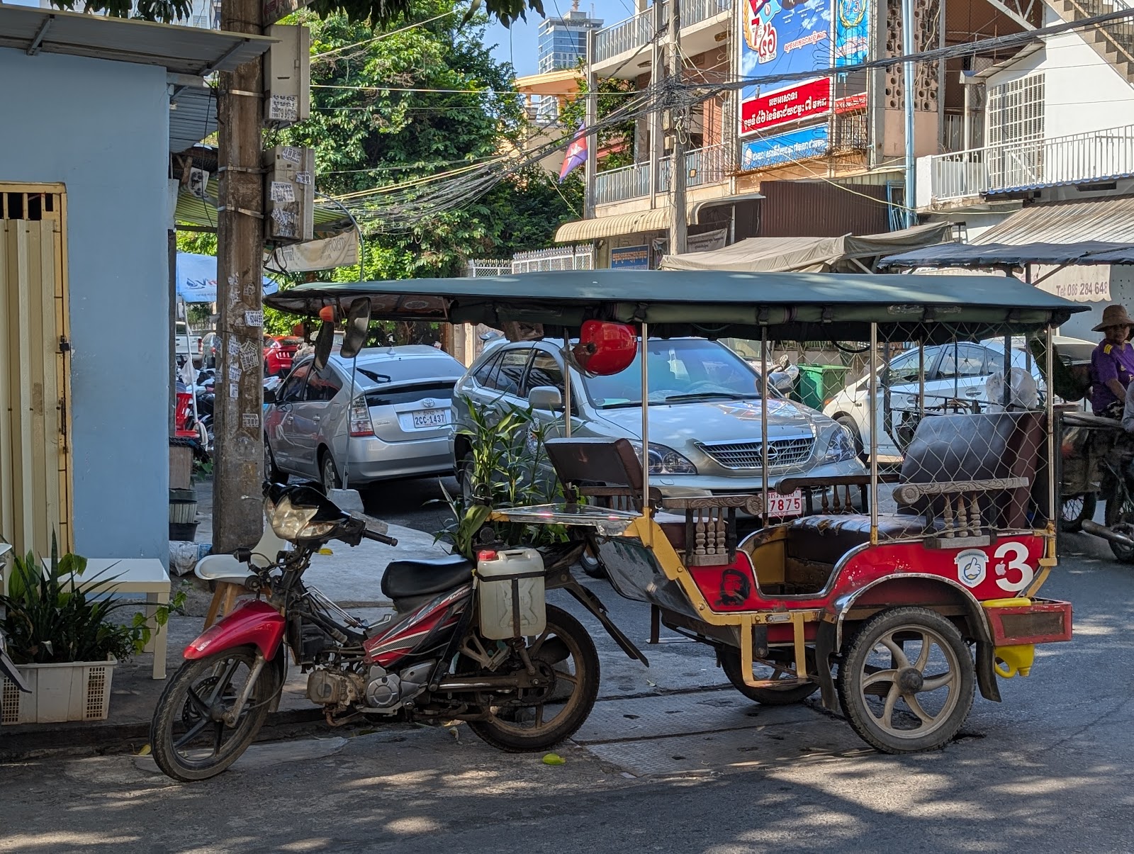 In the foreground, a motorbike attached to a passenger trailer
                 equipped with a canopy. In the background, a street with some
                 parked cars, someone in another motorbike, and a row of
                 apartments. In front of the apartments, cables converge onto
                 a telephone pole