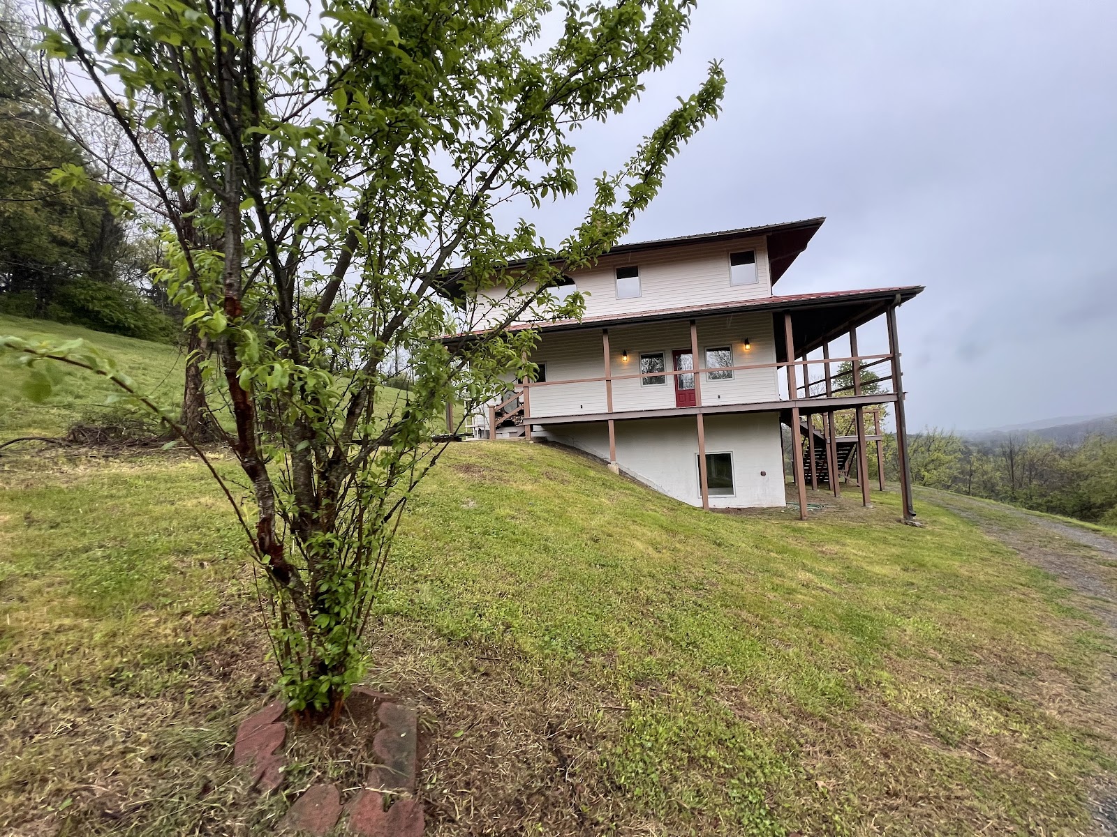Exterior view of a two-story house with light siding and a wooden deck, built on a grassy slope. A small tree stands in the foreground under an overcast sky.